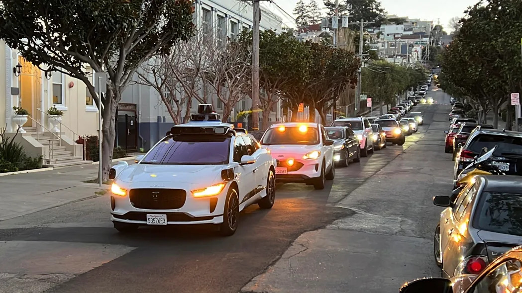 a white car blocks a line of cars waiting behind it on a city street. It's a Waymo self-driving taxi.