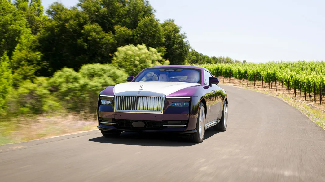 A purple and white Rolls-Royce Spectre car drives down a road with trees in the background.