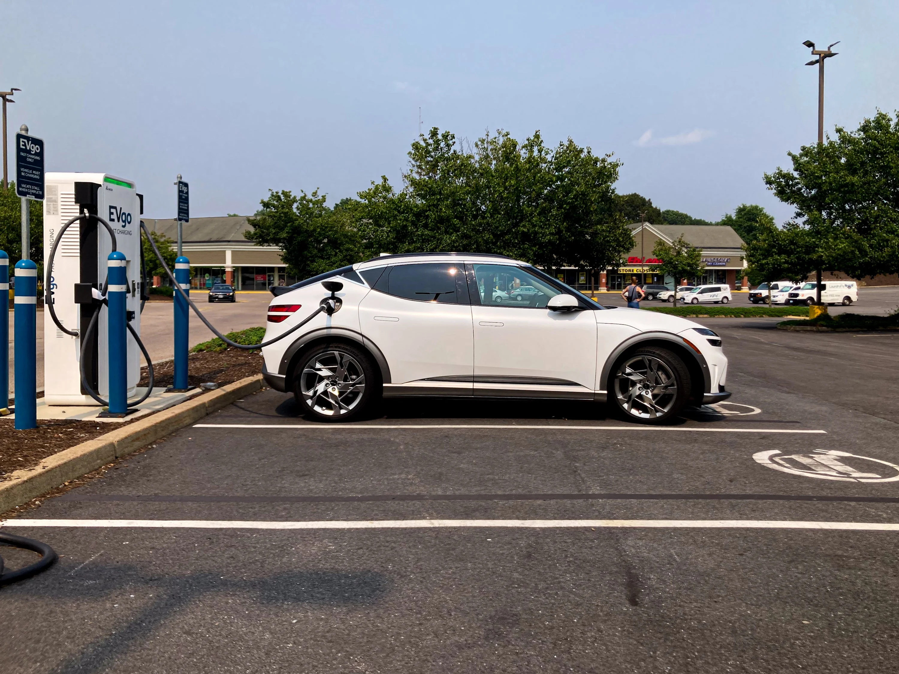 A white Genesis GV60 electric SUV charging at an EVgo station, with trees and sky in the background.