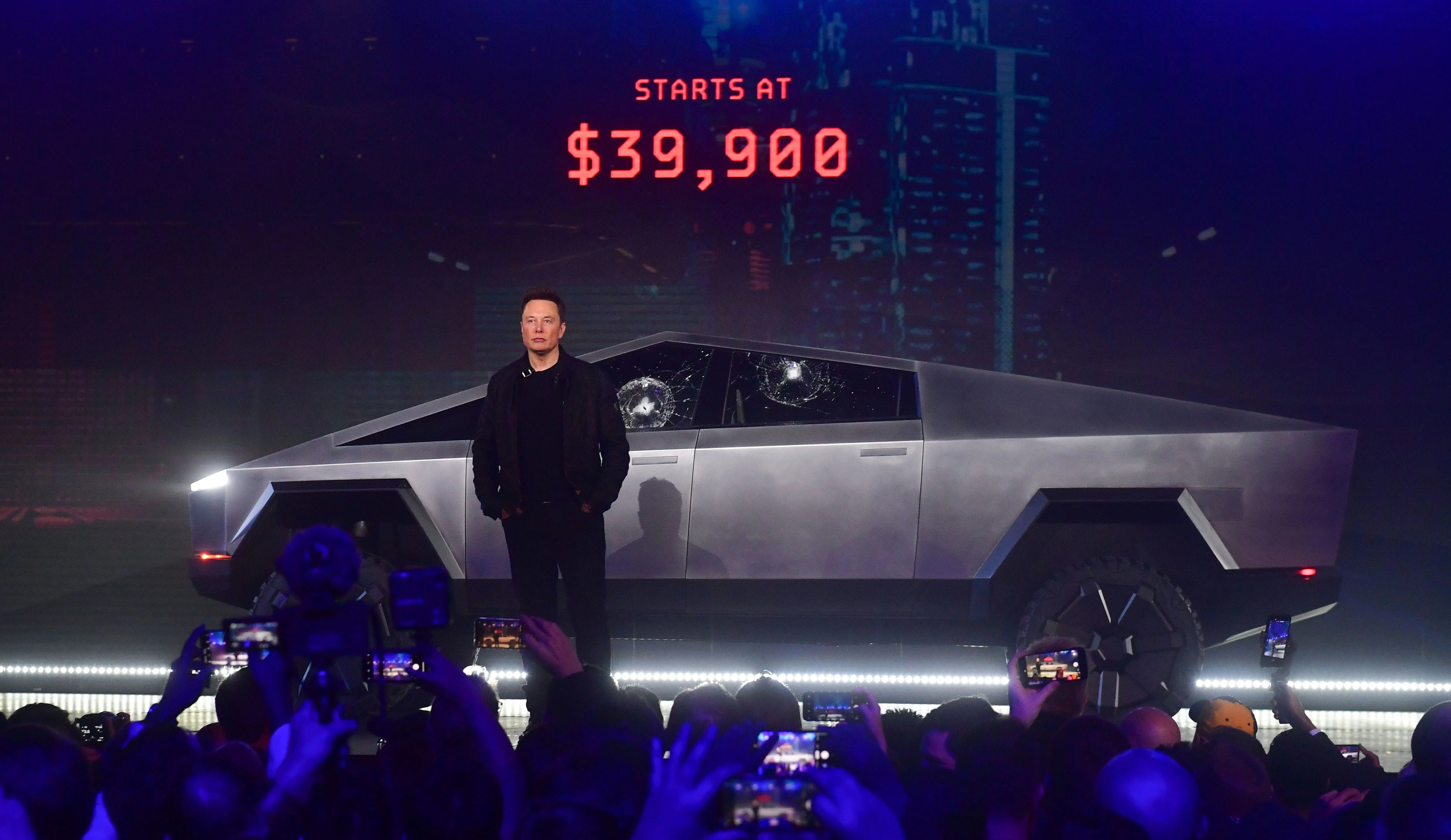 Elon Musk stands in front of Tesla's all-electric Cybertruck on a stage with the price in red lights behind him.