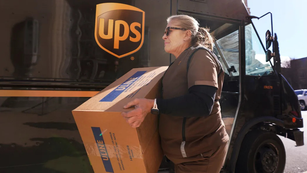 A blond woman in a brown UPS uniform carries a box in front of a brown UPS truck