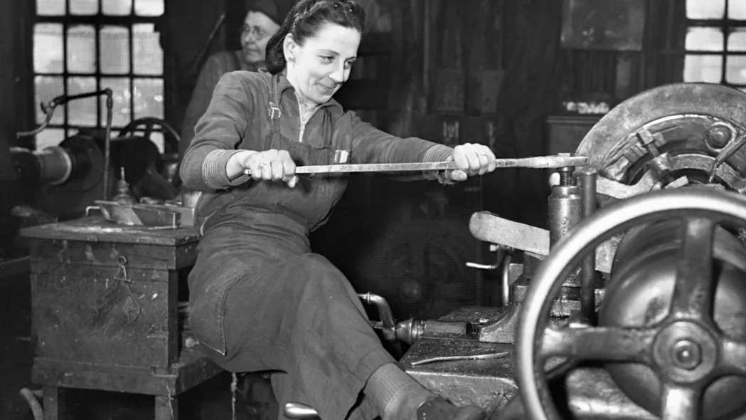 A woman braces with her foot to operate an axle lathe at a car wheel manufacturer in Buffalo, New York. The company began employing women for the war effort when its men went to fight. April, 1943.