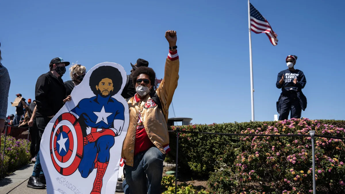 TOPSHOT - A protester kneels with a cardboard cut-out of Colin Kaepernick in Captain America costume in front of a US flag near the Golden Gate Bridge during a demonstration against racism and police brutality in San Francisco, California, on June 6, 2020. - Demonstrations are being held across the US following the death of George Floyd on May 25, 2020, while being arrested in Minneapolis, Minnesota. (Photo by Vivian LIN / AFP) (Photo by VIVIAN LIN/AFP via Getty Images)