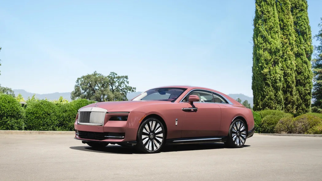 A pink Rolls-Royce Spectre in a parking lot, with blue skies and trees in the background.